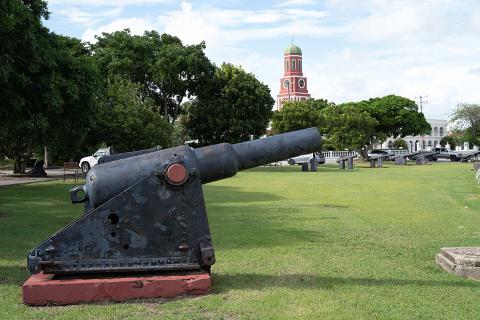This image shows a historical outdoor setting with a large, old cannon in the foreground, mounted on a dark metal carriage with a red wooden base