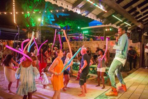 This image shows a vibrant outdoor night event, presumably at a beach or tropical location given the sandy ground and relaxed attire of the attendees