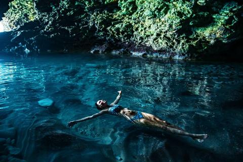 The image depicts a person floating on their back in a crystal-clear water body within a cave-like atmosphere