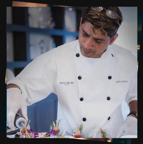 The image shows a chef in a professional kitchen environment, intently focused on preparing or garnishing a dish