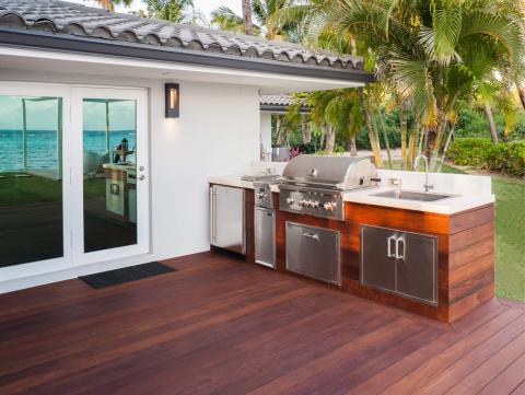 This image depicts an outdoor kitchen setup on a wooden deck with a luxurious tropical backdrop
