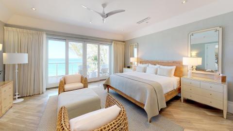 The image shows a well-appointed bedroom with a modern and serene aesthetic, overlooking a beach