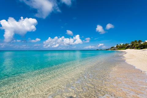 This image captures a serene beach scene with a crystal clear turquoise sea, gentle waves lapping onto a sandy beach, and a blue sky filled with fluffy white clouds