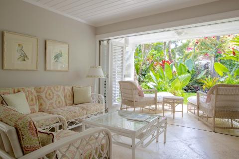 This image shows a bright and airy indoor living room that opens up to a lush tropical garden visible through a large sliding glass door