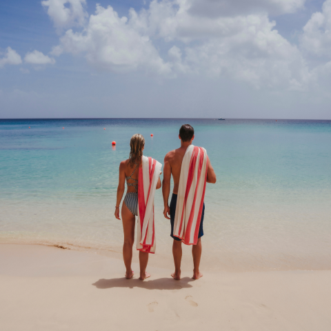 The image shows a man and a woman standing on a beautiful sandy beach, facing the clear turquoise waters of the sea