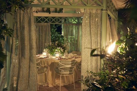 This image shows an elegantly set up dining area in a gazebo-like structure with lush green foliage surrounding it