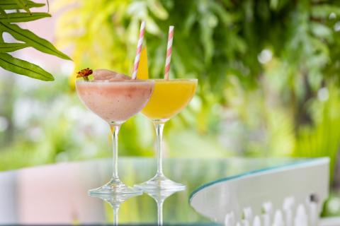 The image features two cocktails placed on a glass table in an outdoor setting surrounded by lush greenery
