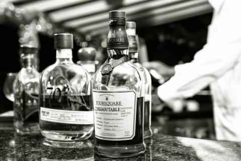 This black and white image captures a selection of liquor bottles on a bar counter, focusing on a specific bottle of Foursquare Redoubtable rum, produced in Barbados