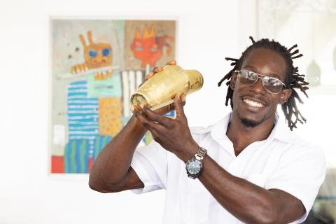 The image shows a cheerful man in a white shirt and glasses, holding a golden cocktail shaker