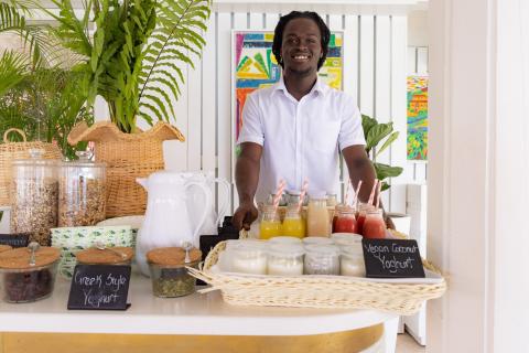 This image features a man standing behind a well-organized refreshment table at what appears to be a resort or similar venue