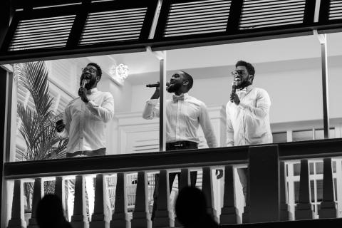 This black and white image captures three men standing on a raised platform, each holding a microphone and passionately singing