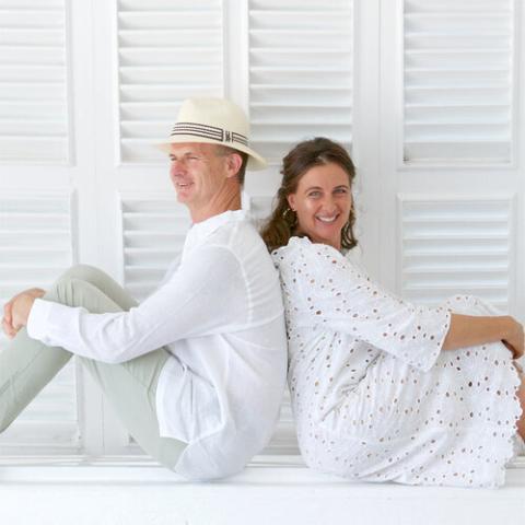 The image shows a man and a woman sitting back-to-back against a white wooden background with horizontal shutters