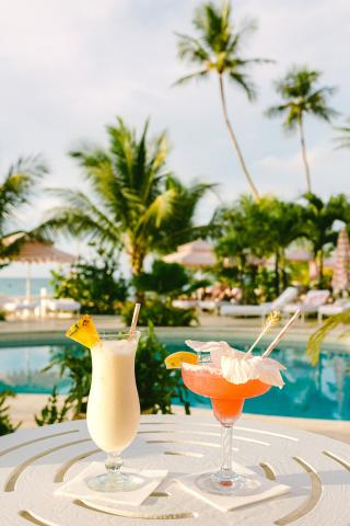 The image shows two tropical cocktails on a white circular table beside a luxurious pool