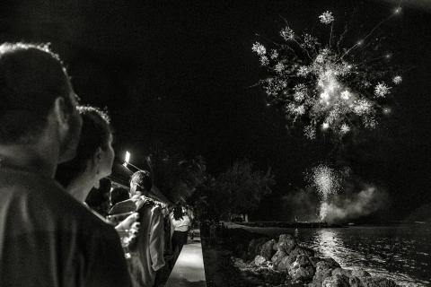 This black and white image captures a group of people watching a fireworks display at night by a waterfront