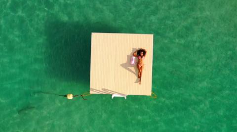 This image shows an aerial view of a woman lying on a floating wooden platform in a clear, turquoise sea