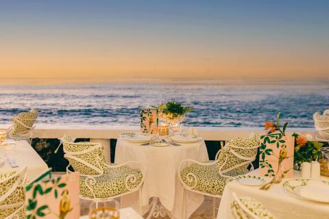 The image features an elegantly set outdoor dining area with a breathtaking view of the sea during what appears to be sunset