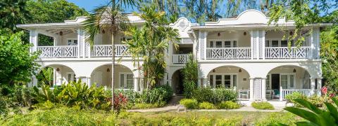 This image displays a beautiful, large two-story colonial-style building surrounded by lush greenery and tropical plants
