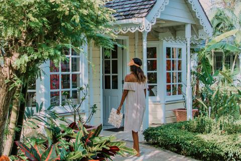 The image shows a woman walking towards a charming cottage surrounded by lush greenery
