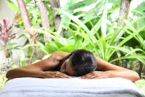 The image shows a woman lying face down on a massage table outdoors, surrounded by lush green plants and trees which create a serene and relaxing atmosphere