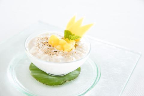 The image shows a bowl of creamy rice pudding garnished with diced mango, a sprig of mint, and what appears to be a decorative piece of mango cut in a crown-like shape