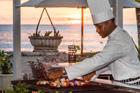 This image shows a chef grilling assorted skewers by the seaside at sunset