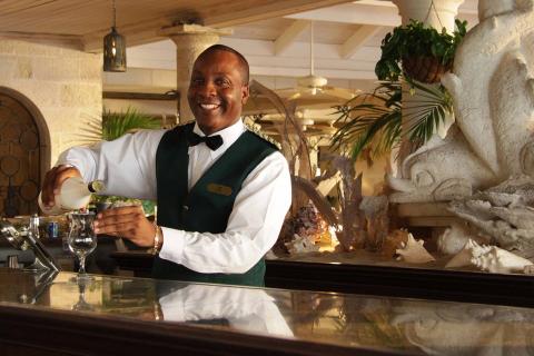 The image features a smiling bartender, dressed in an elegant green and white uniform with a black bowtie