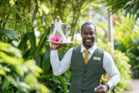 This image shows a man dressed in a professional uniform consisting of a vest and tie, smiling pleasantly as he holds a tray