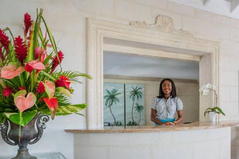 This image shows a woman standing at a reception desk, set in a warmly lit space designed with tropical and elegant elements