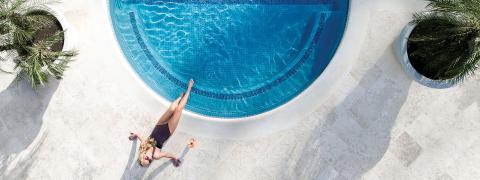 The image features an aerial view of a luxurious swimming pool with a curved design, showcasing sparkling blue water surrounded by a smooth stone deck