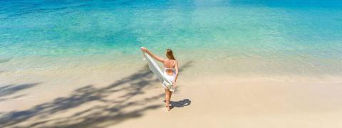 The image shows a person standing on a pristine sandy beach, facing the crystal clear waters of the ocean
