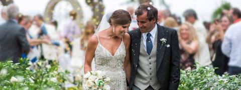 The image shows a newlywed couple walking down what appears to be an aisle, surrounded by guests and decorations at their wedding ceremony