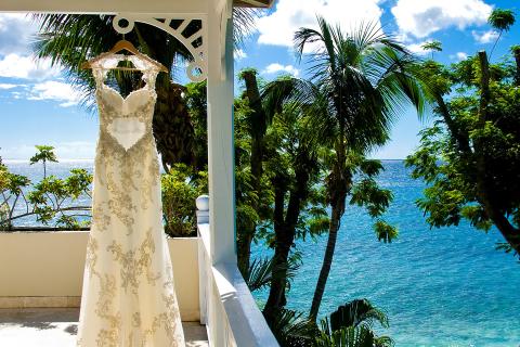 This image features a beautiful wedding dress hanging from a white pavilion, set against a scenic backdrop of lush tropical trees and a clear blue sea