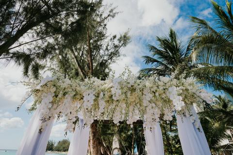 This image shows an elegant floral arch setup for what seems to be an outdoor wedding or event, located on a sunny beach setting