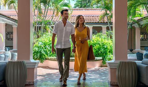 The image features a man and a woman walking through a breezy, elegantly designed arcade or walkway with columns