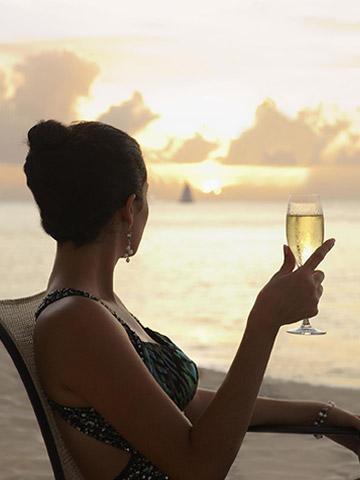 The image shows a woman sitting on a beach chair at sunset, holding a glass of sparkling wine