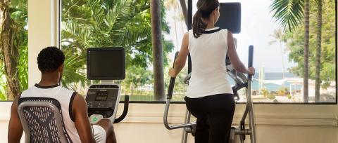 The image shows two people exercising on stationary fitness machines in a gym with a scenic view