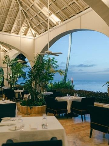 This image shows a sophisticated restaurant setting featuring an elegant design with an open archway offering a view of the ocean
