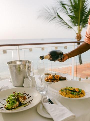The image shows a dining setup on a balcony or an outdoor terrace with a view of the sea and a palm tree