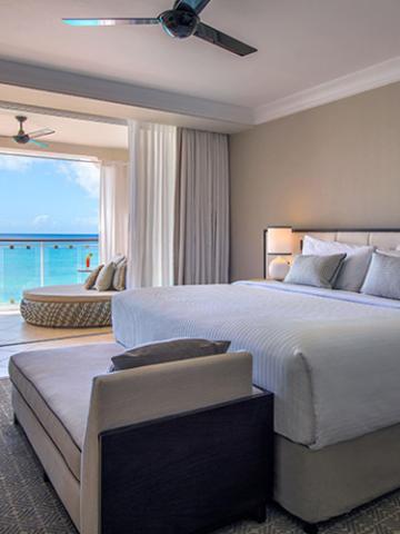 This image features a well-appointed hotel room with a large bed in the foreground, covered in a white bedspread and accompanied by several pillows