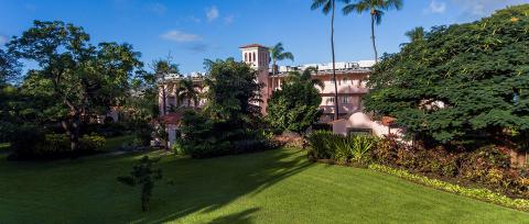 The image displays a sprawling, lush garden in the foreground with a well-manicured lawn, surrounded by various types of trees and flowering shrubs