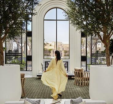 This image shows an elegantly dressed woman in a flowing yellow dress standing in front of a large arched window that offers a view of outdoor scenery