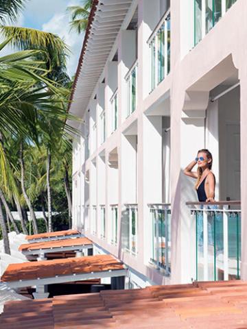 The image shows a woman standing on a balcony of a modern building with a tropical setting