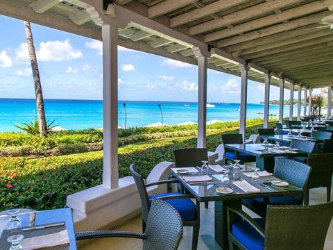 This image shows an outdoor dining area under a covered patio with a beautiful view of a tropical beach