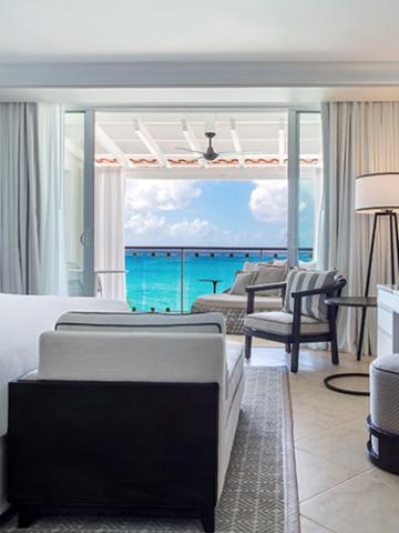 The image depicts a beautifully appointed hotel or resort room with a large window providing a picturesque view of the ocean