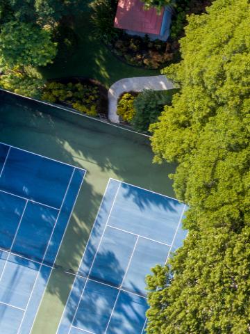 The image shows an aerial view of a blue tennis court alongside a swimming pool, surrounded by lush green trees