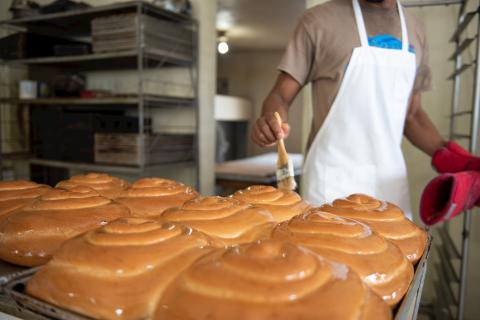 In the image, there is a person in a bakery who appears to be glazing large, freshly baked bread loaves