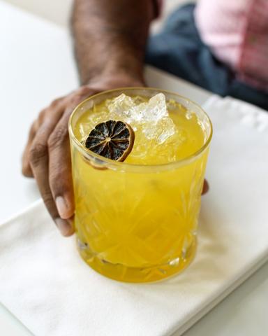 The image shows a hand holding a glass of chilled yellow beverage, topped with ice and a dried citrus slice, possibly lime or lemon