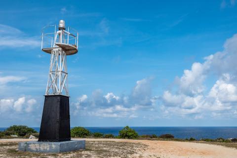 The image features a metal structure, which appears to be a small lighthouse or navigational beacon, set against a serene coastal landscape with clear skies and scattered clouds