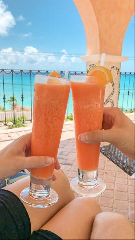 The image shows two people holding glasses of a frothy, orange-colored drink, possibly a cocktail or a fruit smoothie, garnished with slices of orange