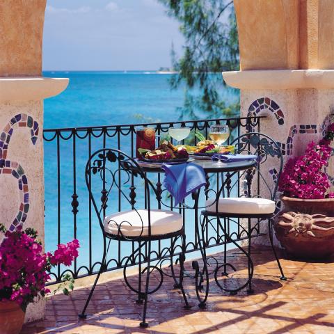 The image depicts a picturesque seaside balcony setup for a meal